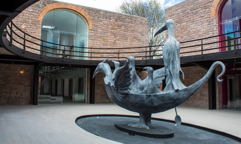 Escultura Cocodrilo de Leonora Carrington en bronce instalada en un patio arquitectónico, con figuras fantásticas y lenguaje surrealista.