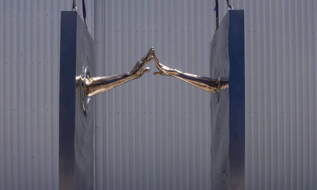 Escultura MAKTUB It Is Written de Lorenzo Quinn con brazos en bronce pulido emergiendo de placas de acero inoxidable.