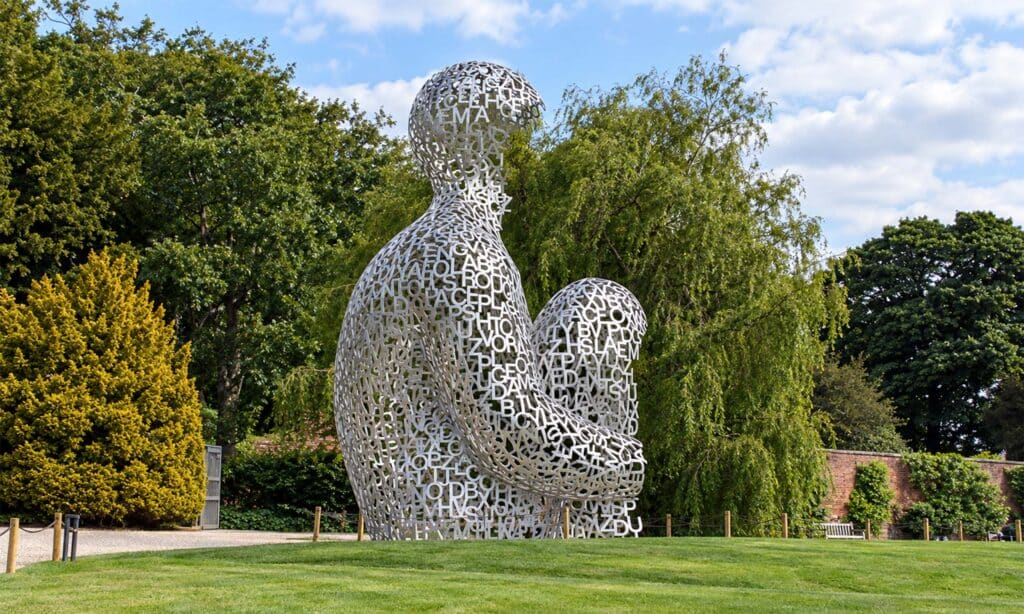 Escultura de Jaume Plensa con dos figuras humanas sentadas, formadas por letras metálicas y situadas al aire libre en un jardín.